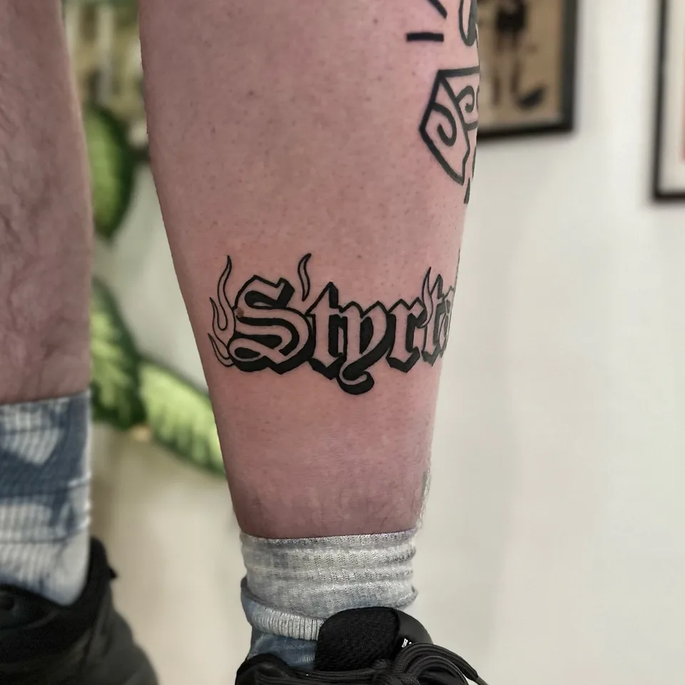 Tatuaż z grubym konturem – słowo w stylu gotyckim z motywem płomieni na łydce — Tattoo, Wujek Darek Tattoo, Warszawa